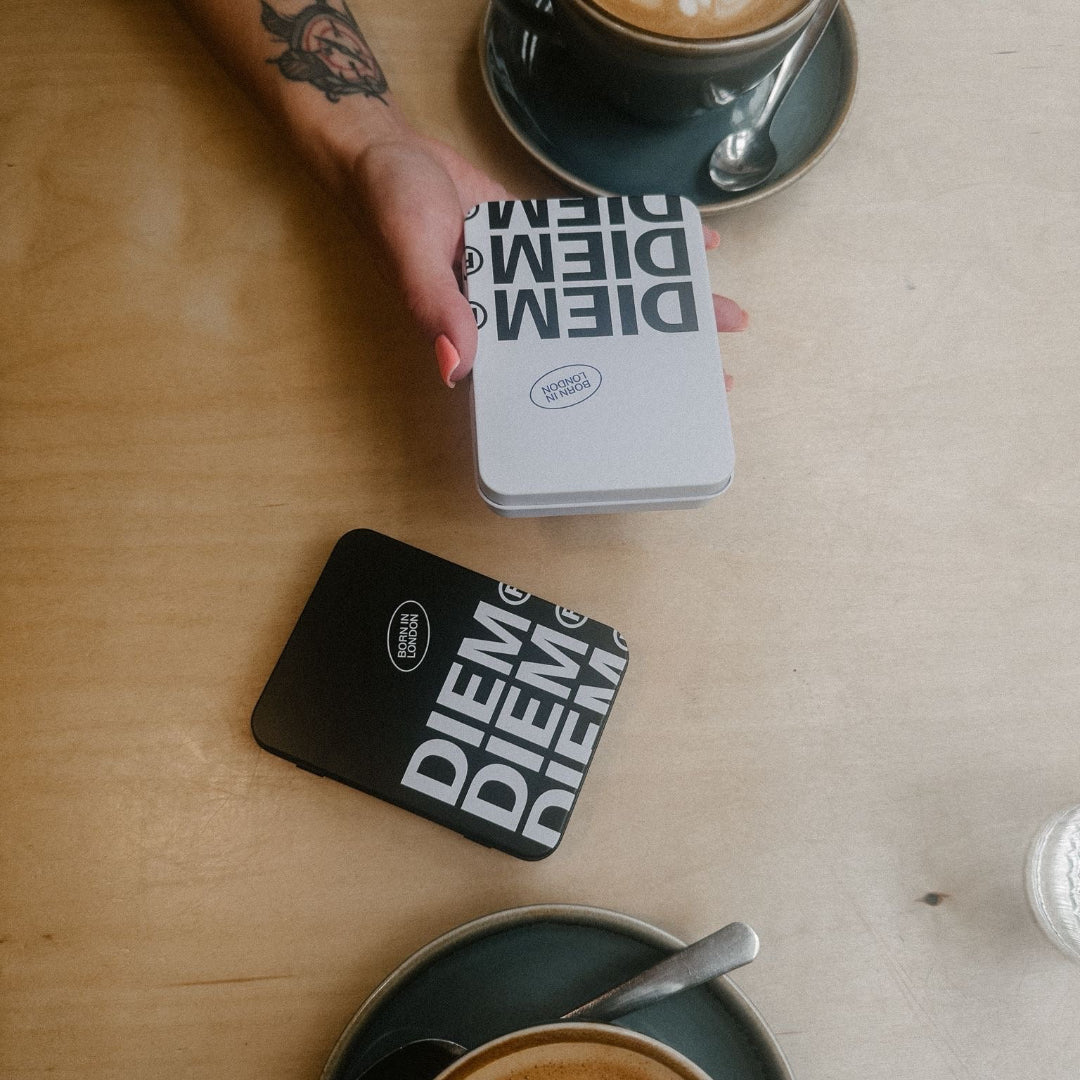 Two cups of coffee with latte art on a table, one with a hand holding a fragrance discovery tin 'DIEM'.